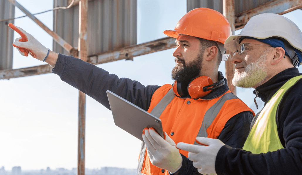 Two construction workers in hard hats and hi-vis vests pointing at a structure and holding a tablet.