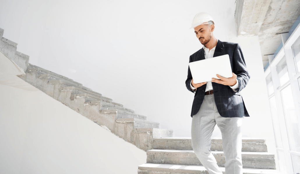 Construction professional in a hard hat reviewing plans on a laptop while standing on an unfinished concrete staircase