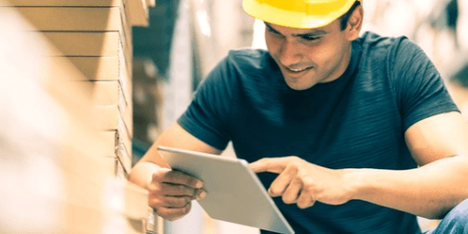 Worker in a yellow hard hat checking a tablet beside stacked materials in a warehouse