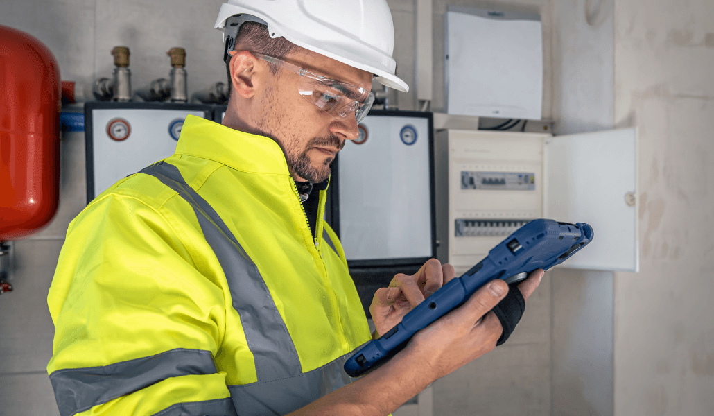 Maintenance worker in hard hat and safety glasses using a rugged tablet to inspect building mechanical and electrical systems.