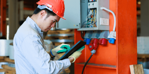 Industrial electrician inspecting electrical panel and using a digital tablet.