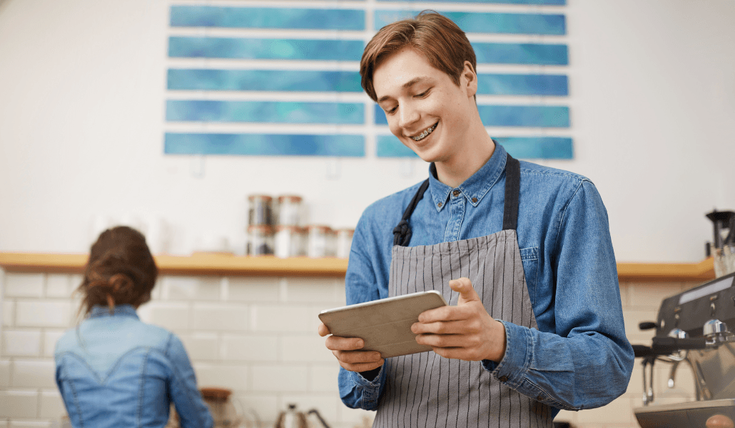 A young cafe worker in an apron smiles while using a tablet, with a colleague working in the background.