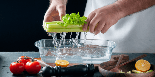 Someone rinsing celery under running water over a glass bowl, with tomatoes and lemon nearby.