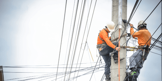 Two utility workers wearing safety harnesses and orange shirts climb a power pole to repair electrical wires.